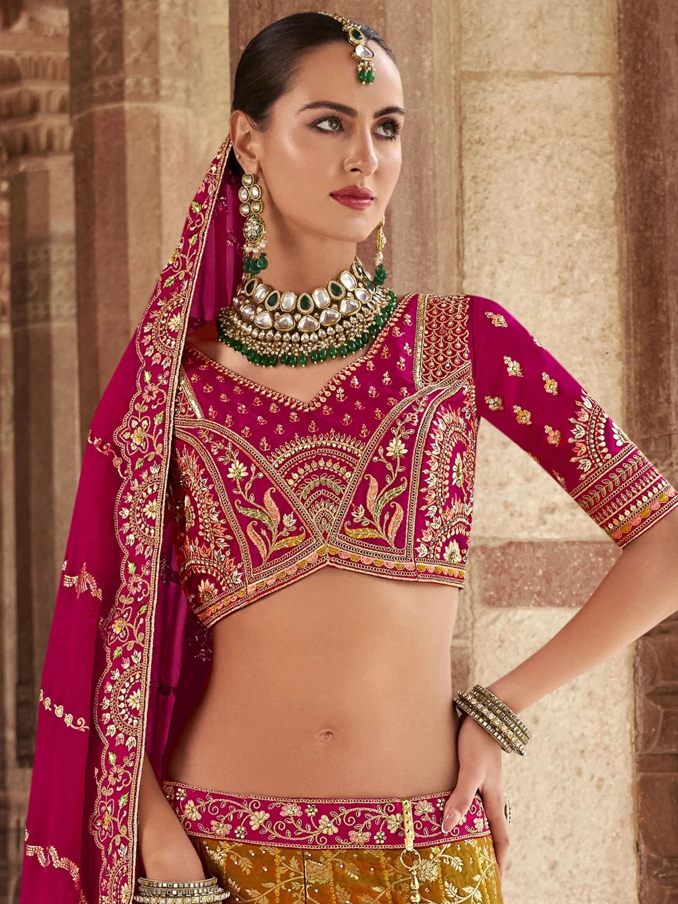 Woman in pink embroidered lehenga choli with gold detailing and green jewelry, traditional Indian ethnic wear