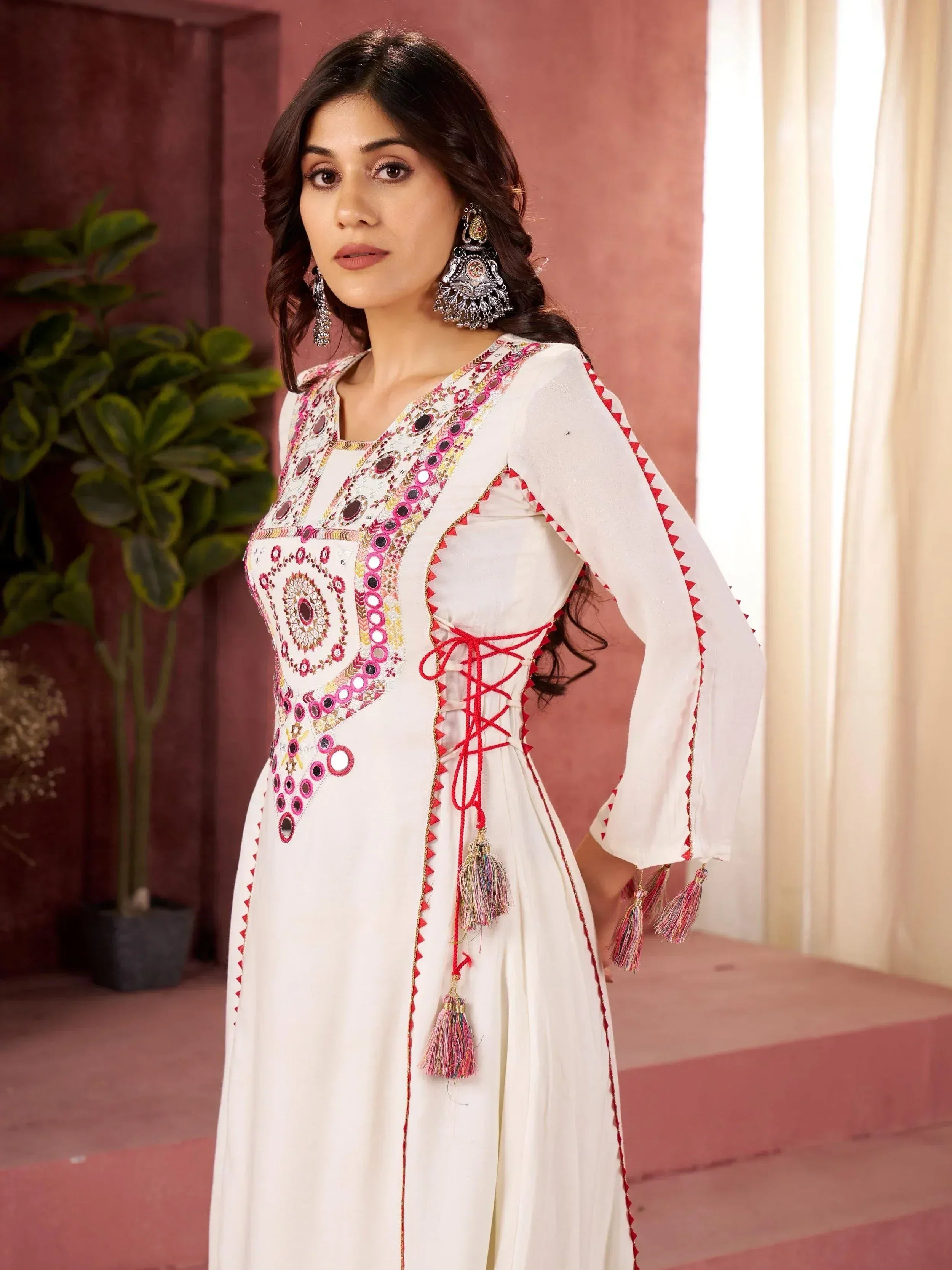 White ethnic gown with mirror work, red lace-up back, and tassels, styled with jhumka earrings.