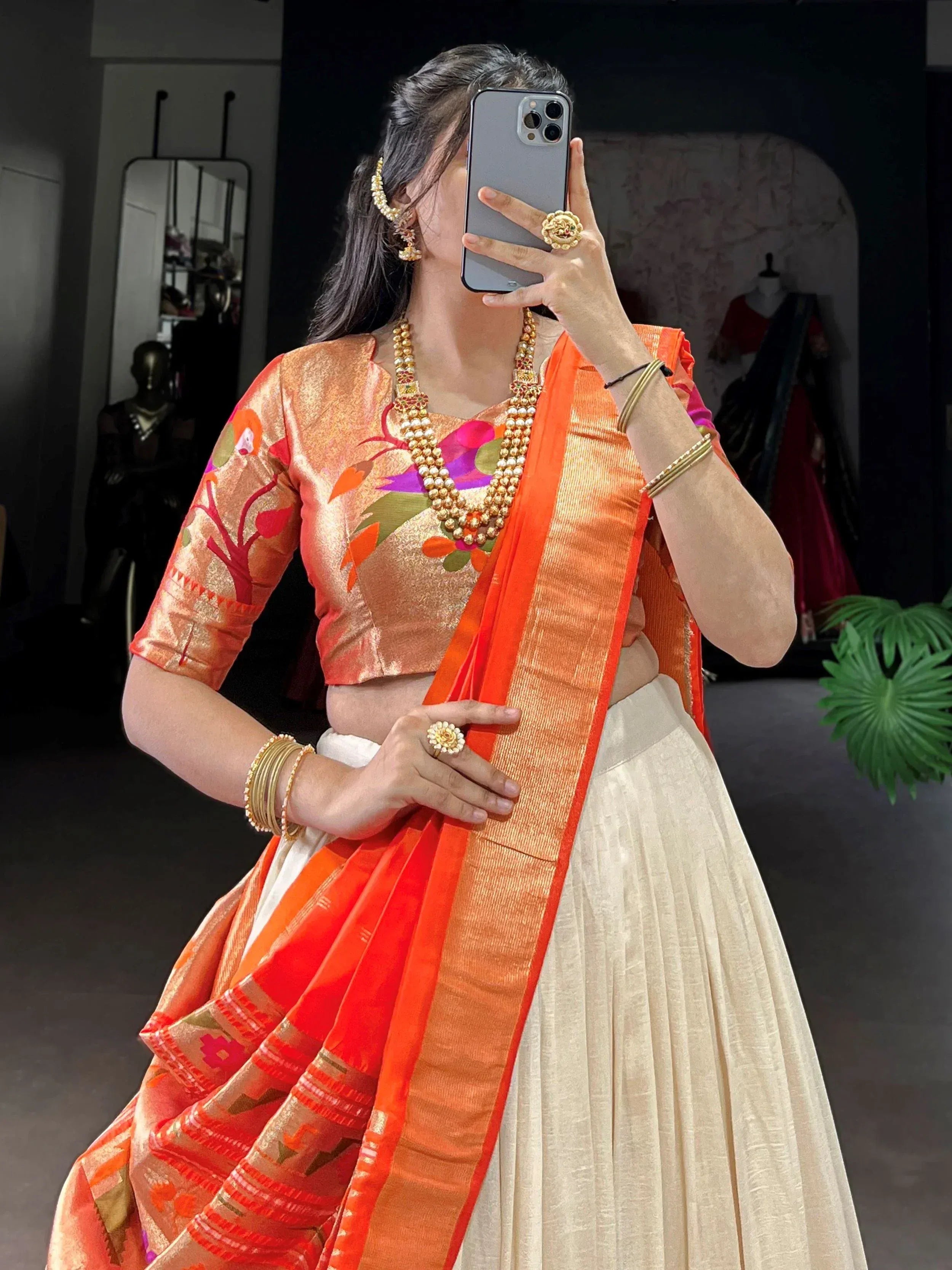 Woman in vibrant orange and cream designer lehenga choli with floral blouse, traditional jewelry, taking mirror selfie