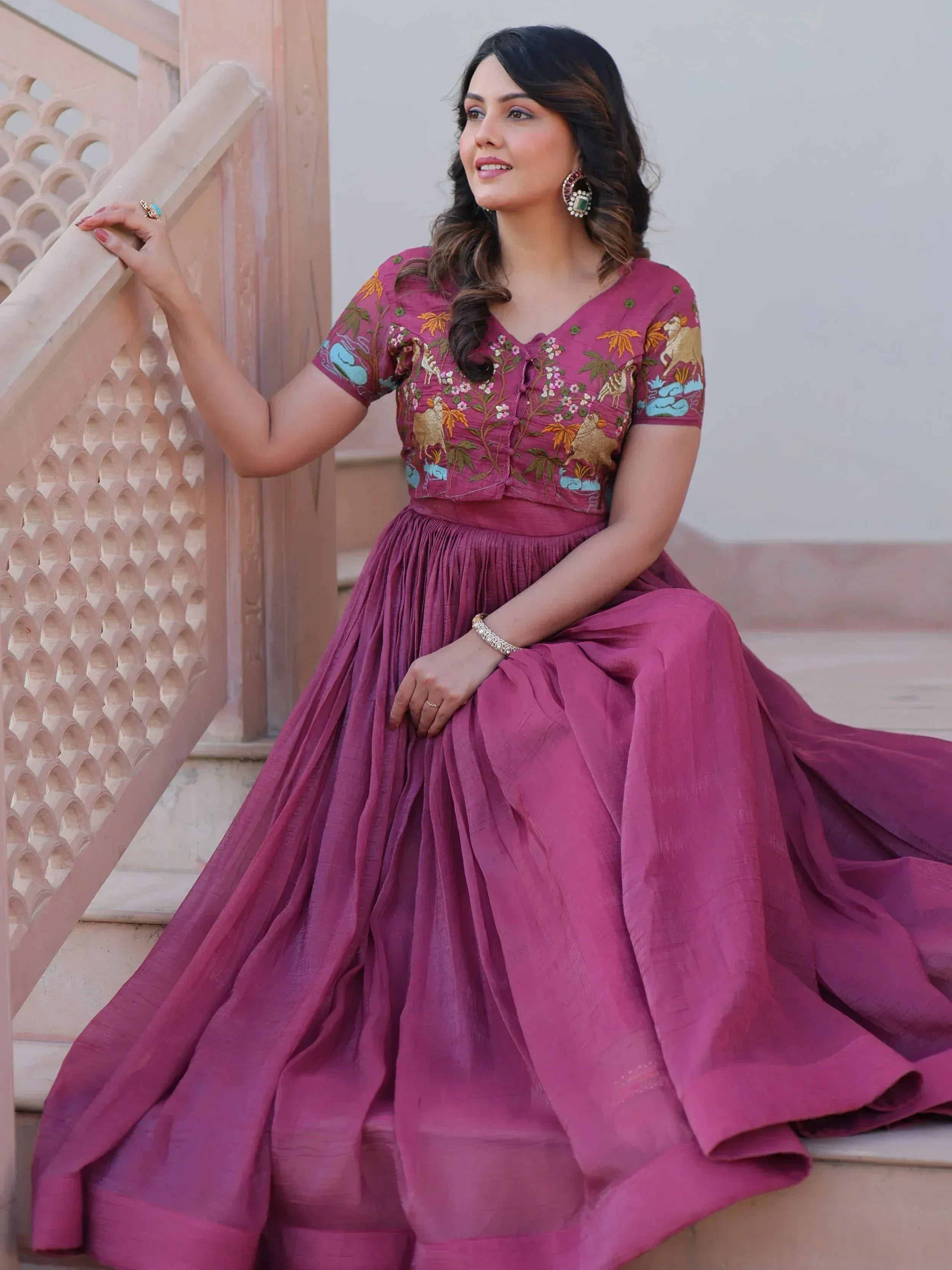 Woman in maroon embroidered ethnic gown with floral and animal motifs, sitting on stairs
