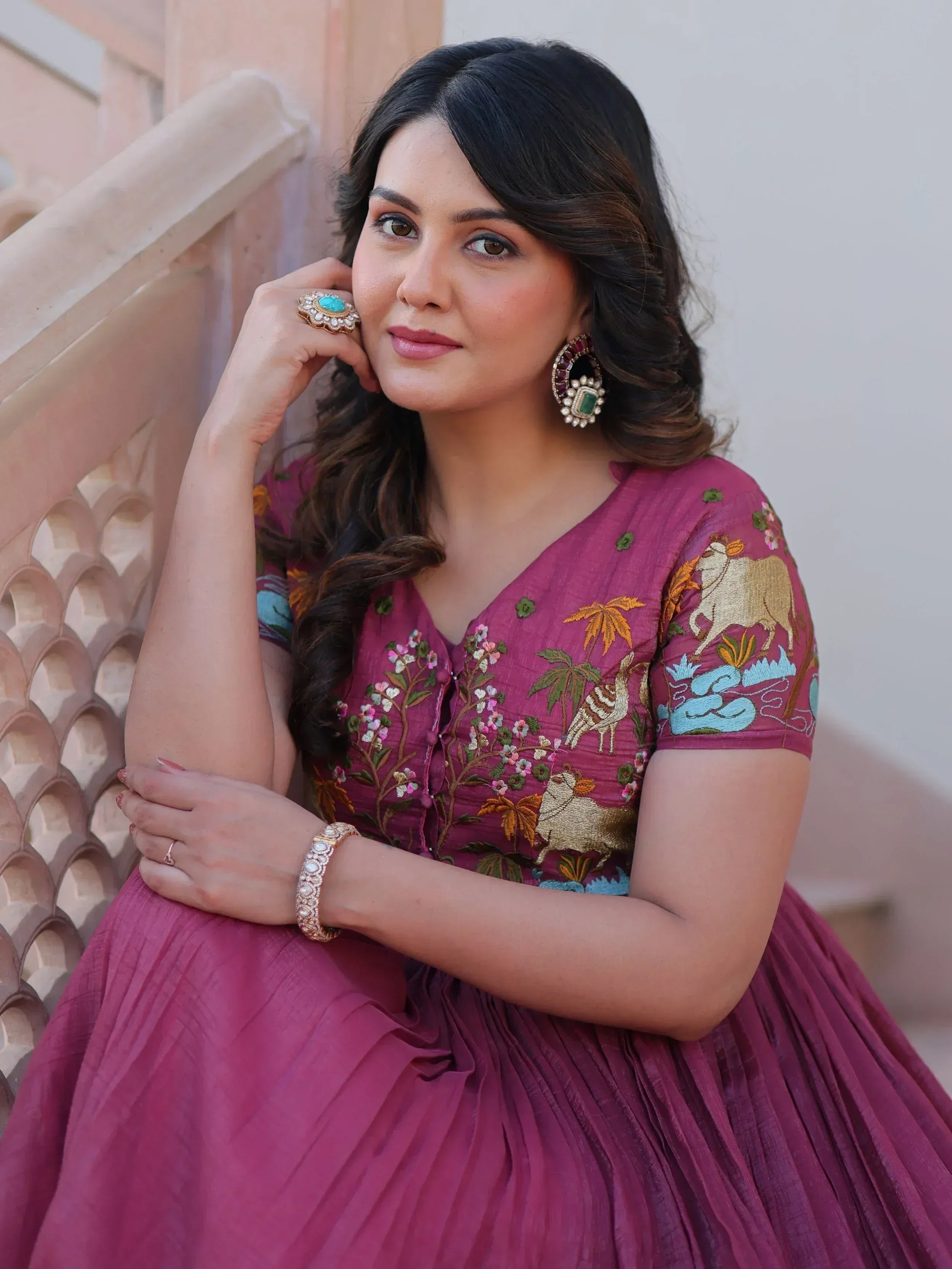 Woman in maroon embroidered ethnic gown with floral and animal motifs, paired with statement jewelry.