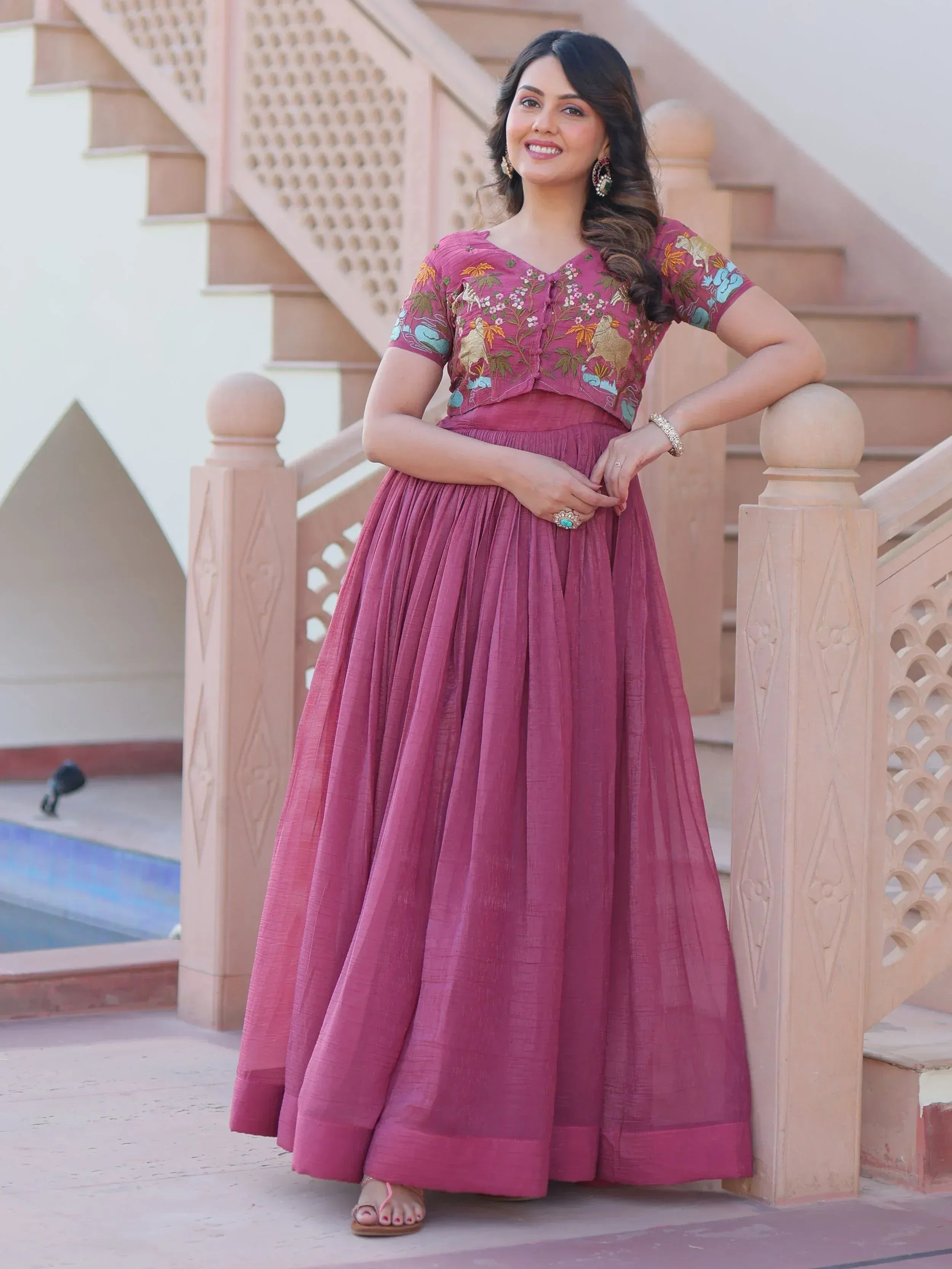 Woman in pink embroidered ethnic gown with floral and animal motifs, standing by staircase