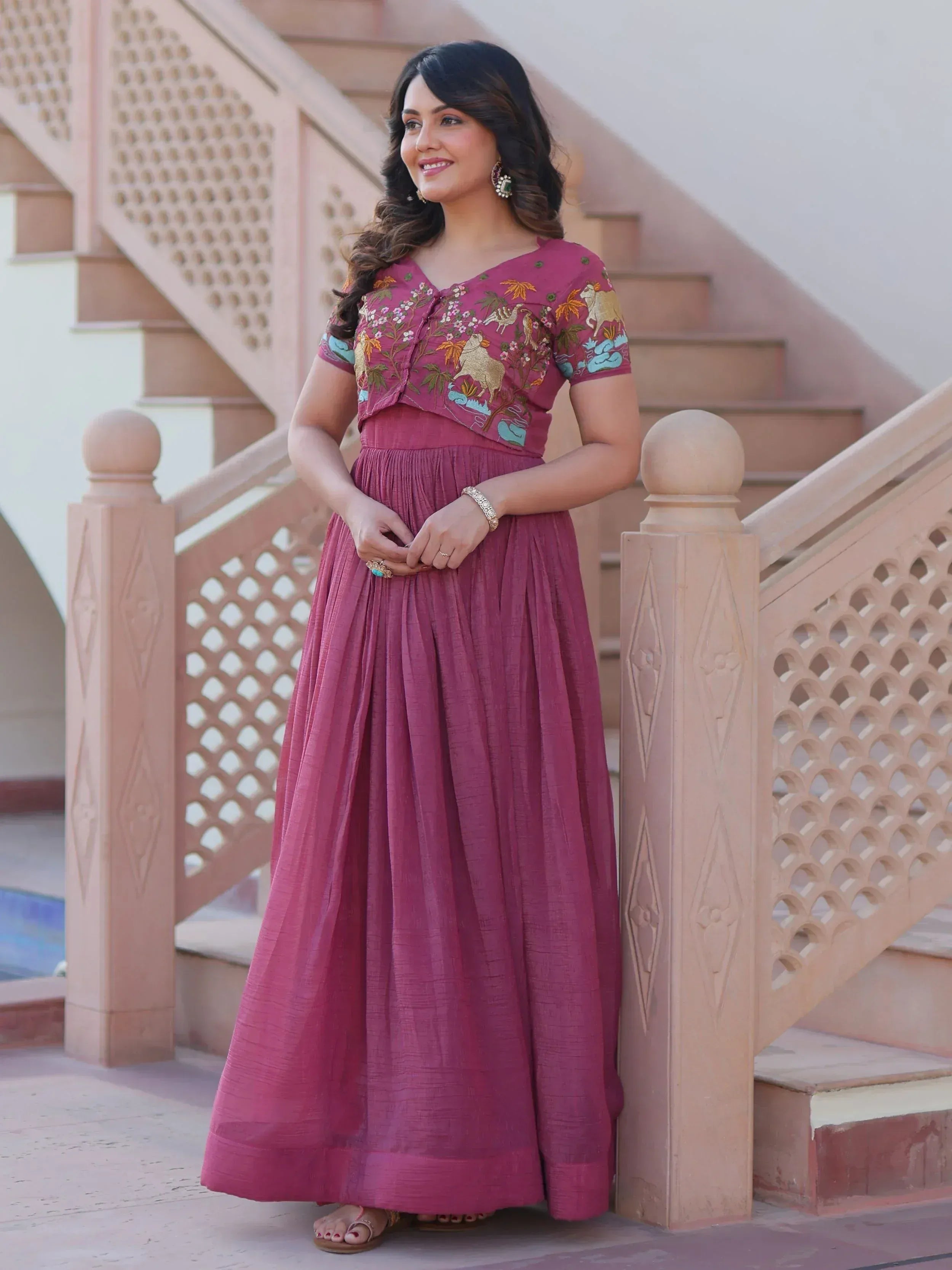 Woman in maroon embroidered ethnic gown with floral and animal motifs, standing by ornate staircase