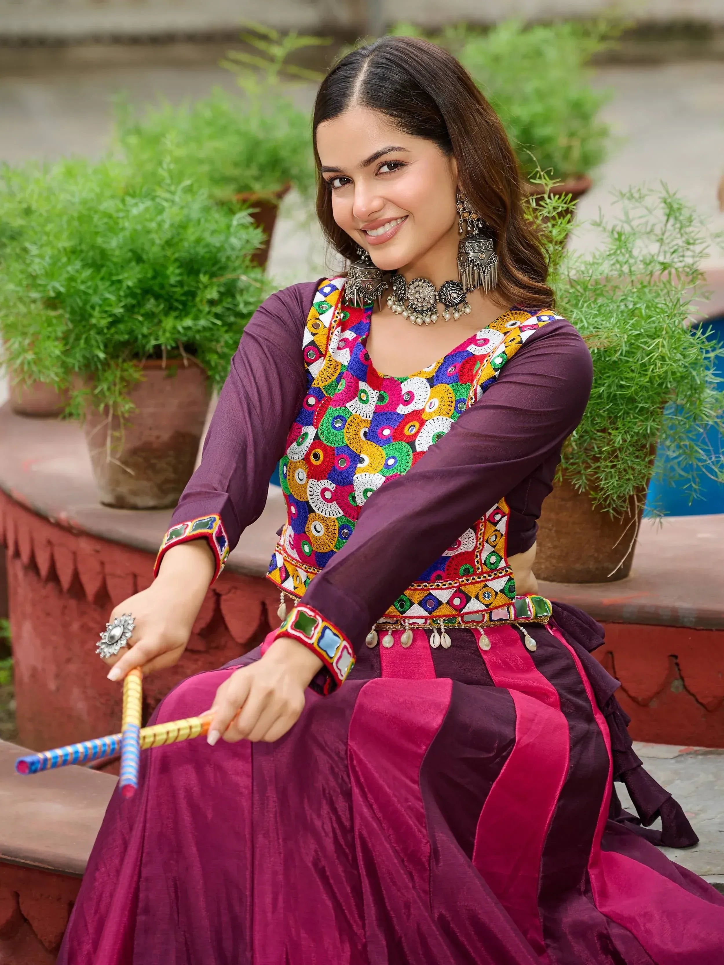 Woman in colorful embroidered ethnic wear with mirror work, holding decorative sticks, outdoor setting
