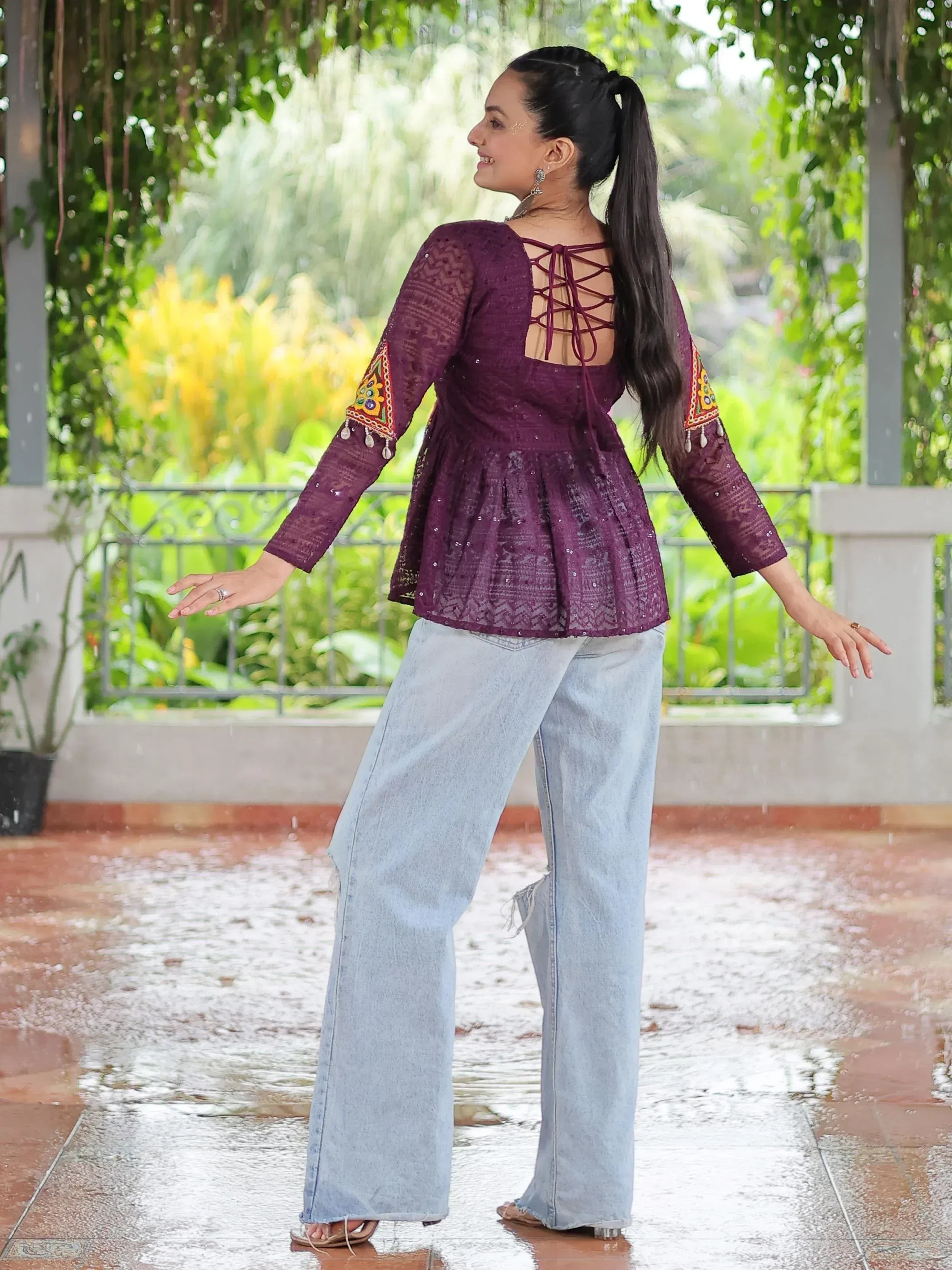 Woman in purple embroidered ethnic top with lace-up back, paired with wide-leg jeans, standing in rain