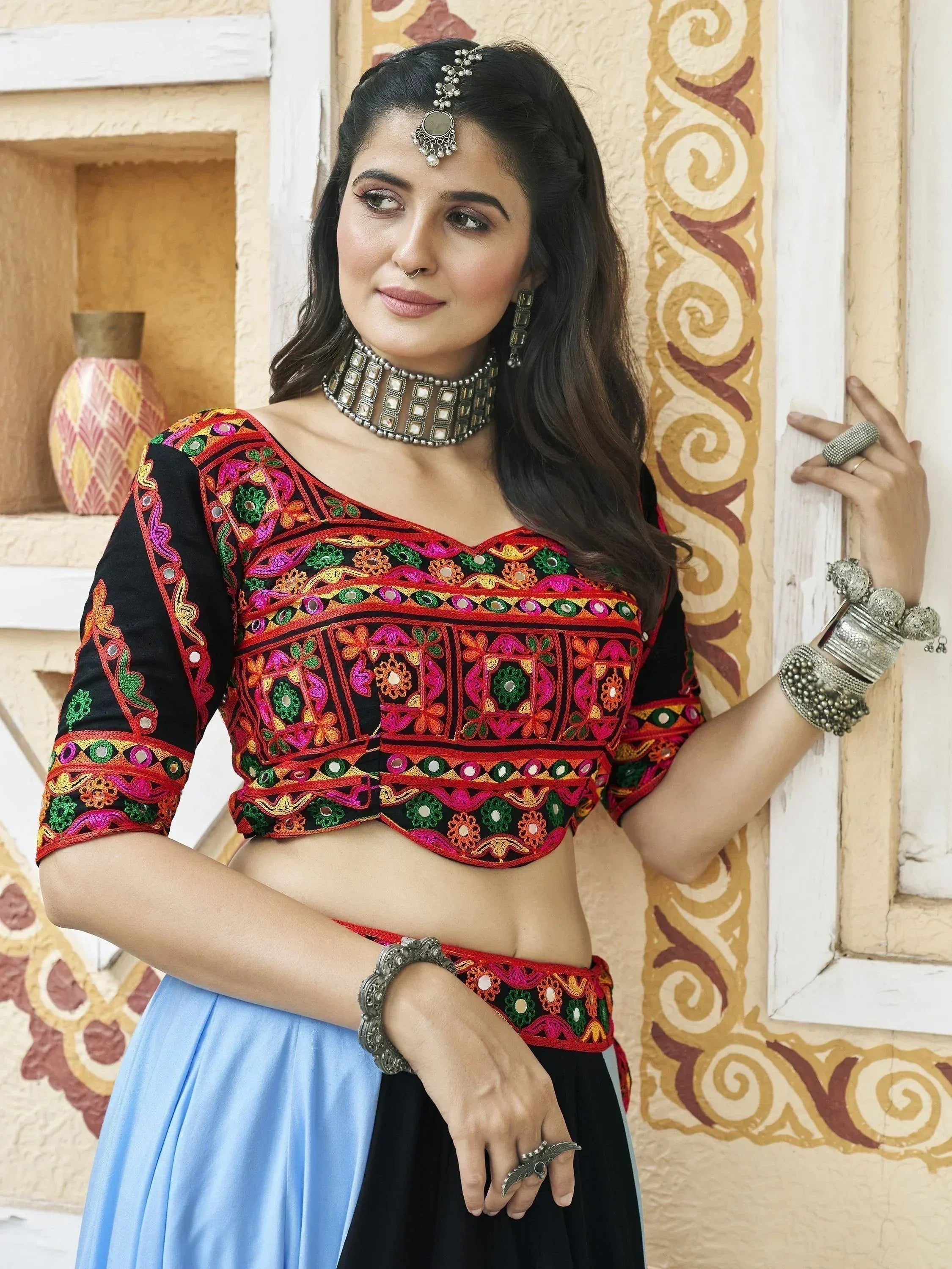 Woman in colorful embroidered Indian ethnic blouse with mirror work, paired with blue and black skirt, traditional jewelry.
