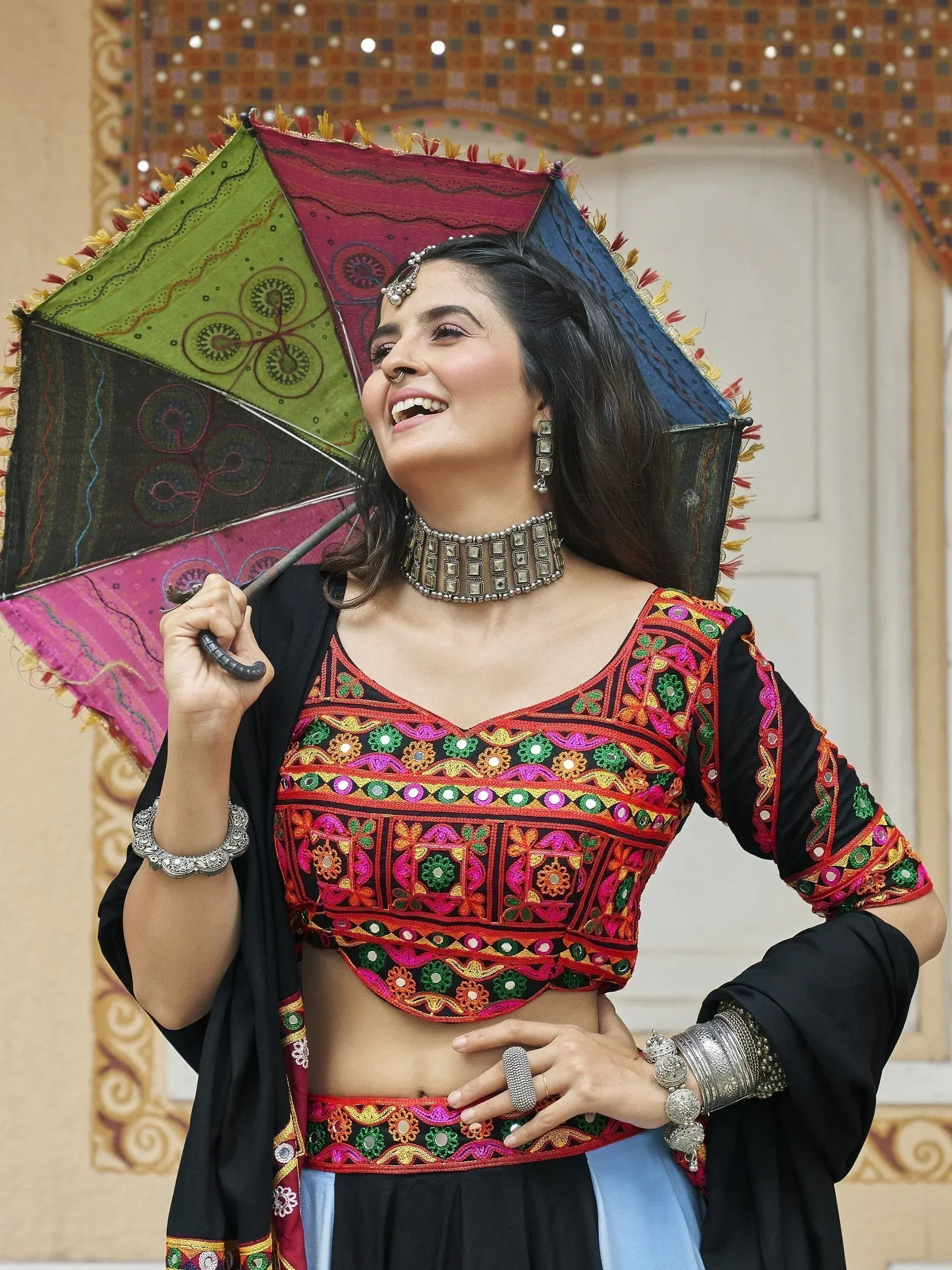 Woman in colorful embroidered Indian ethnic wear with mirror work, holding decorative umbrella, traditional jewelry