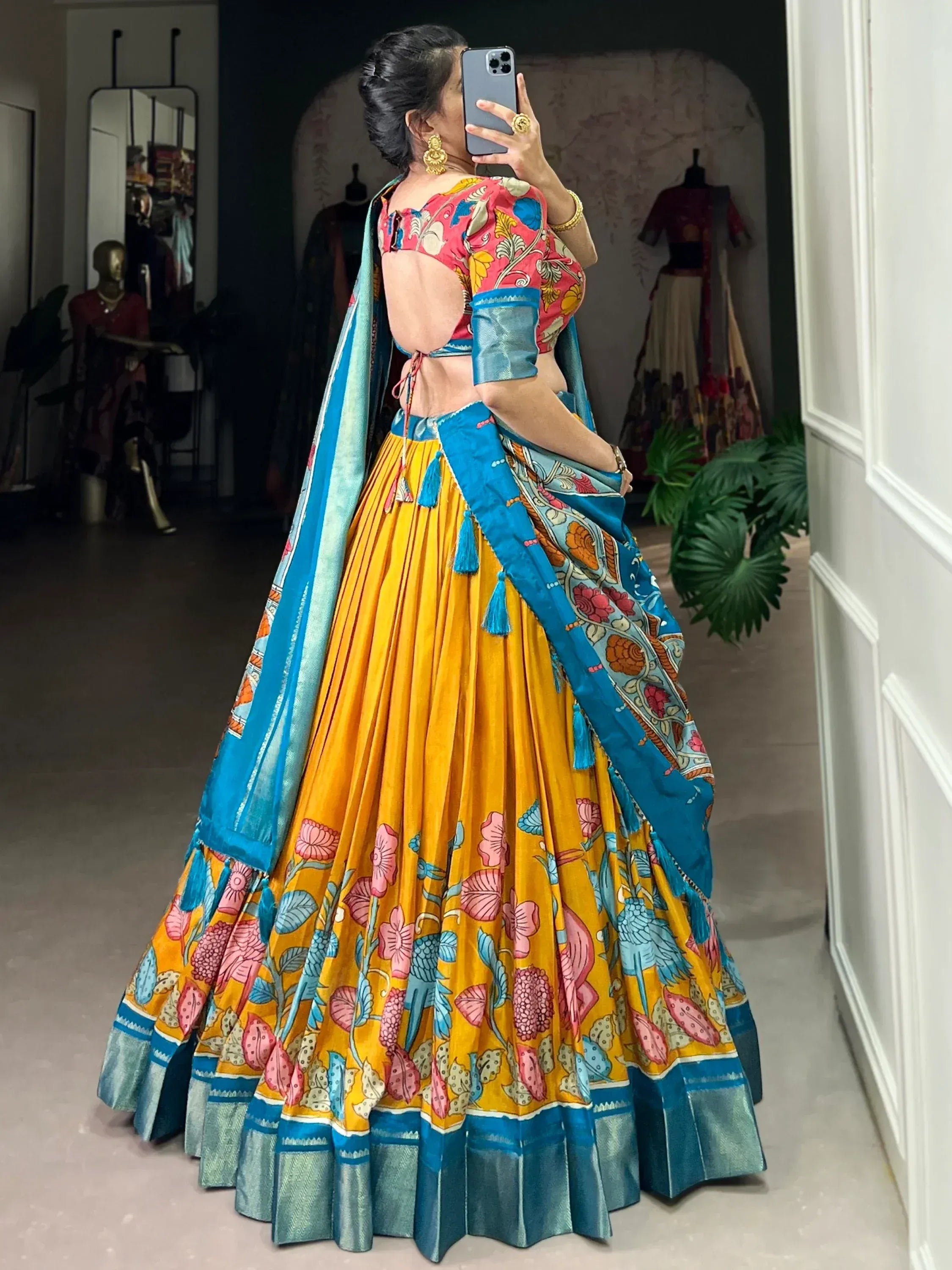 Woman in vibrant yellow floral lehenga choli with blue dupatta, taking mirror selfie in boutique
