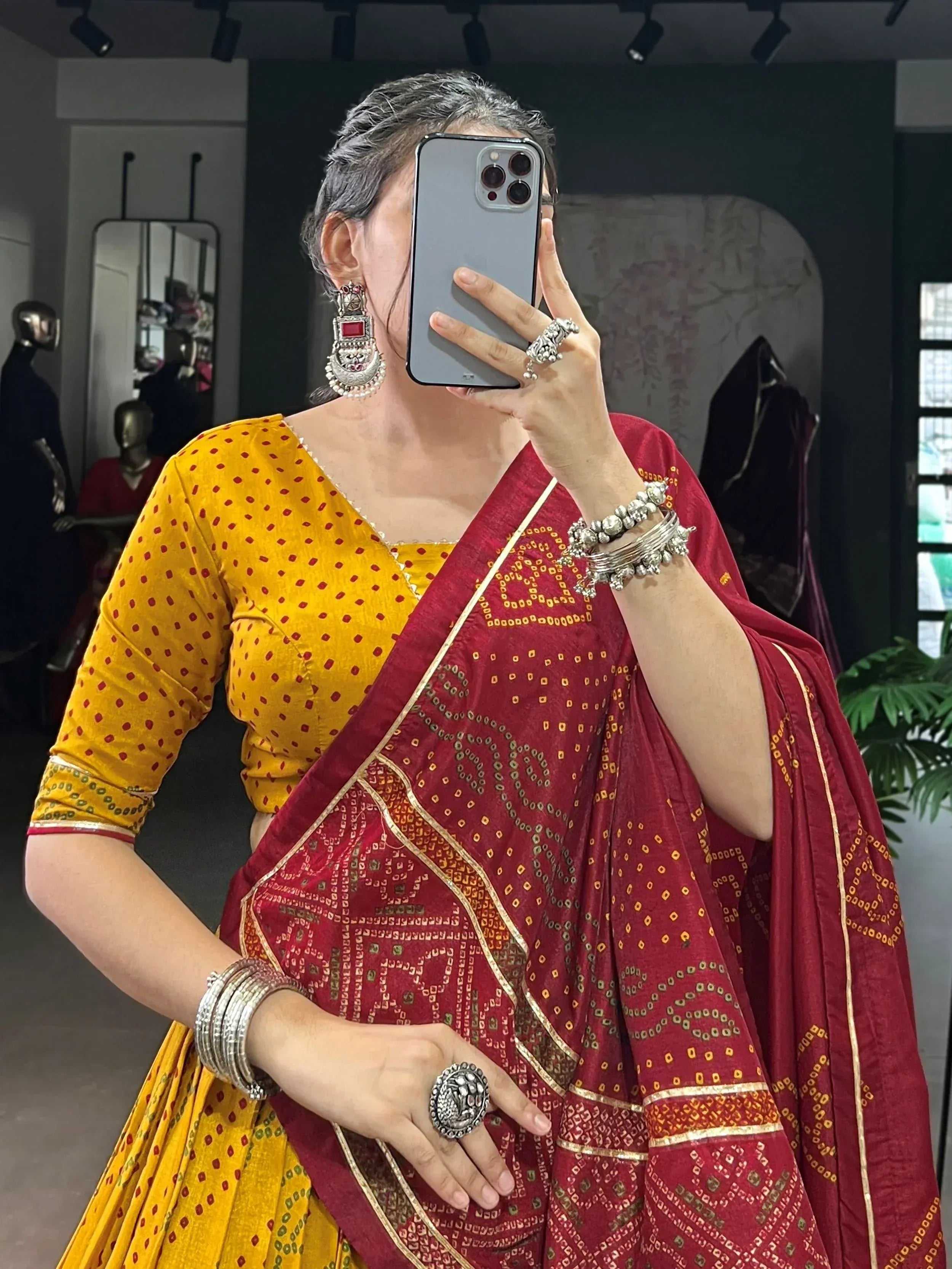 Woman in maroon and yellow ethnic saree with traditional jewelry, taking mirror selfie in boutique