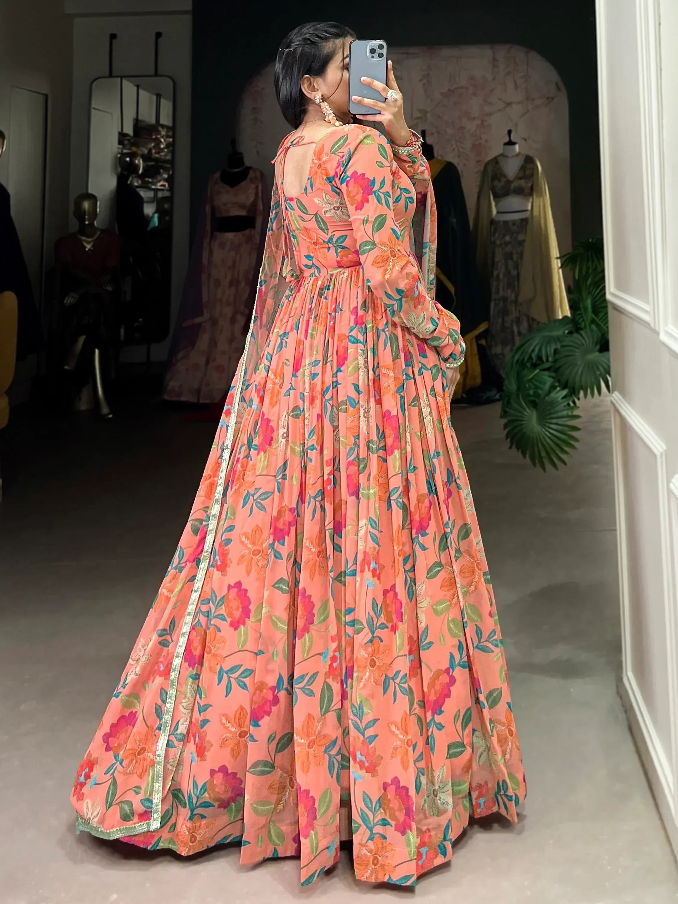 Woman in peach floral anarkali gown with back cutout, taking mirror selfie in boutique