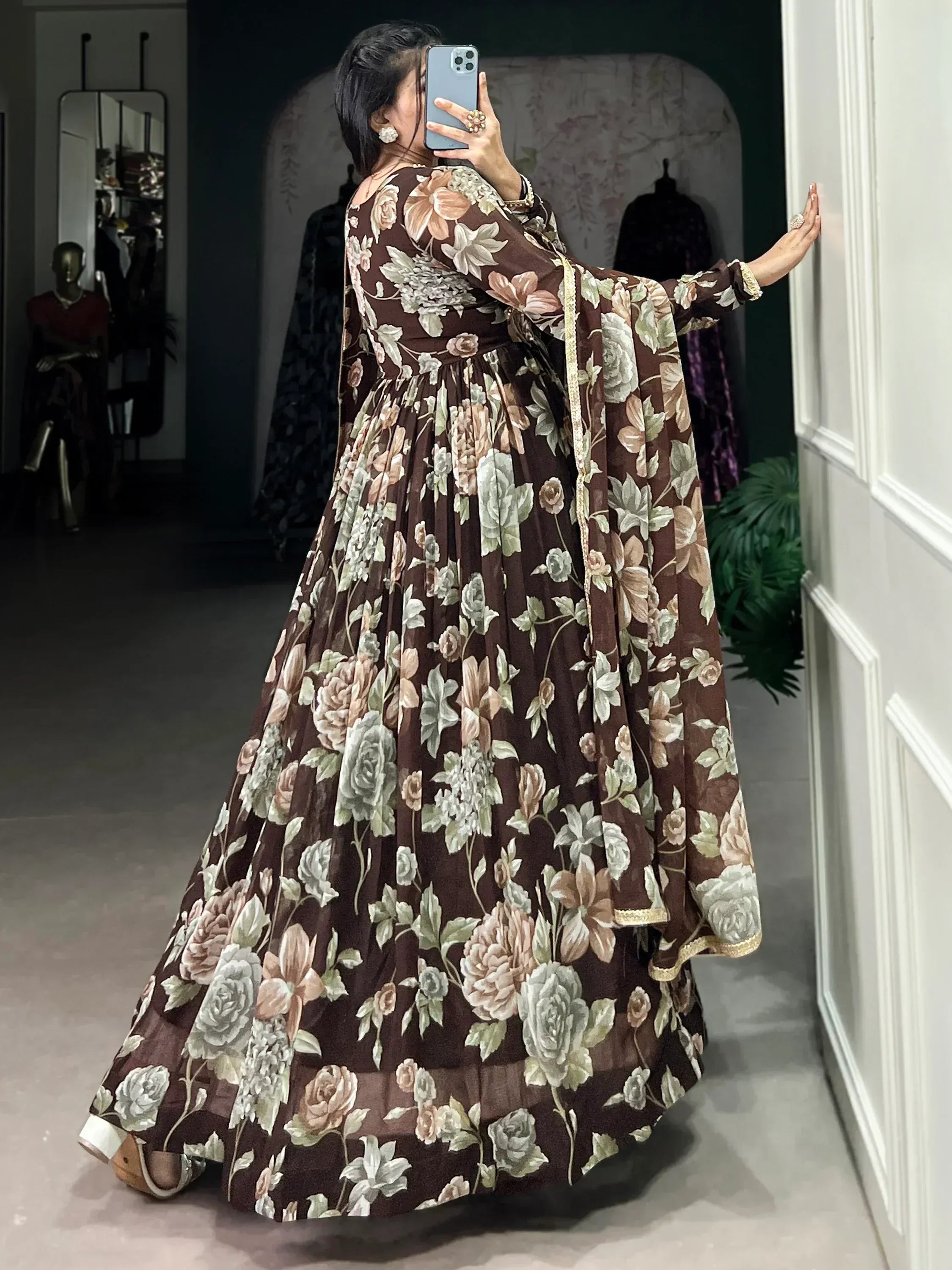 Woman in brown floral print Indian ethnic gown with gold trim, taking mirror selfie in boutique