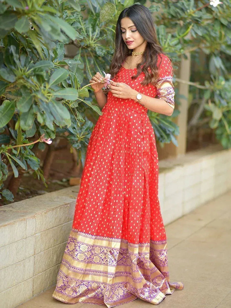 Woman in red ethnic gown with gold motifs and purple brocade border, outdoors near plants