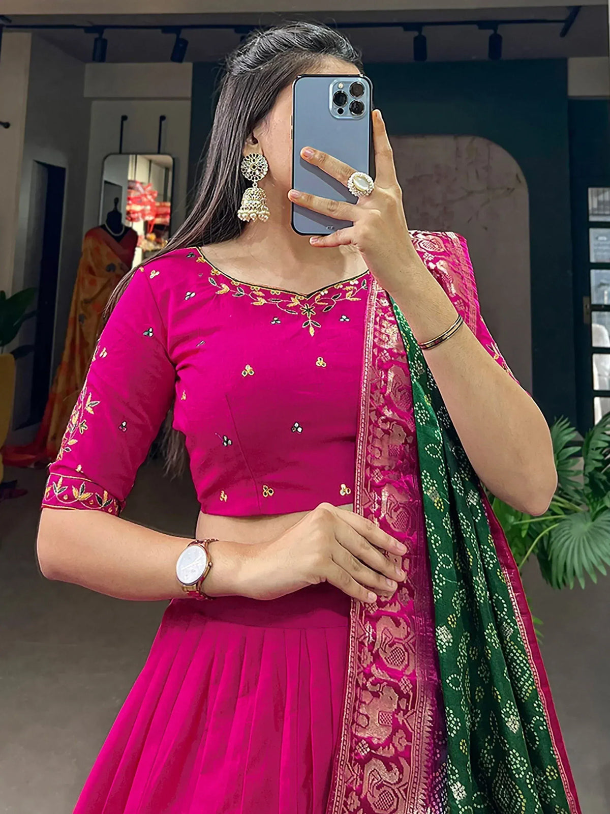 Woman in vibrant pink embroidered lehenga choli with green dupatta, taking mirror selfie in boutique