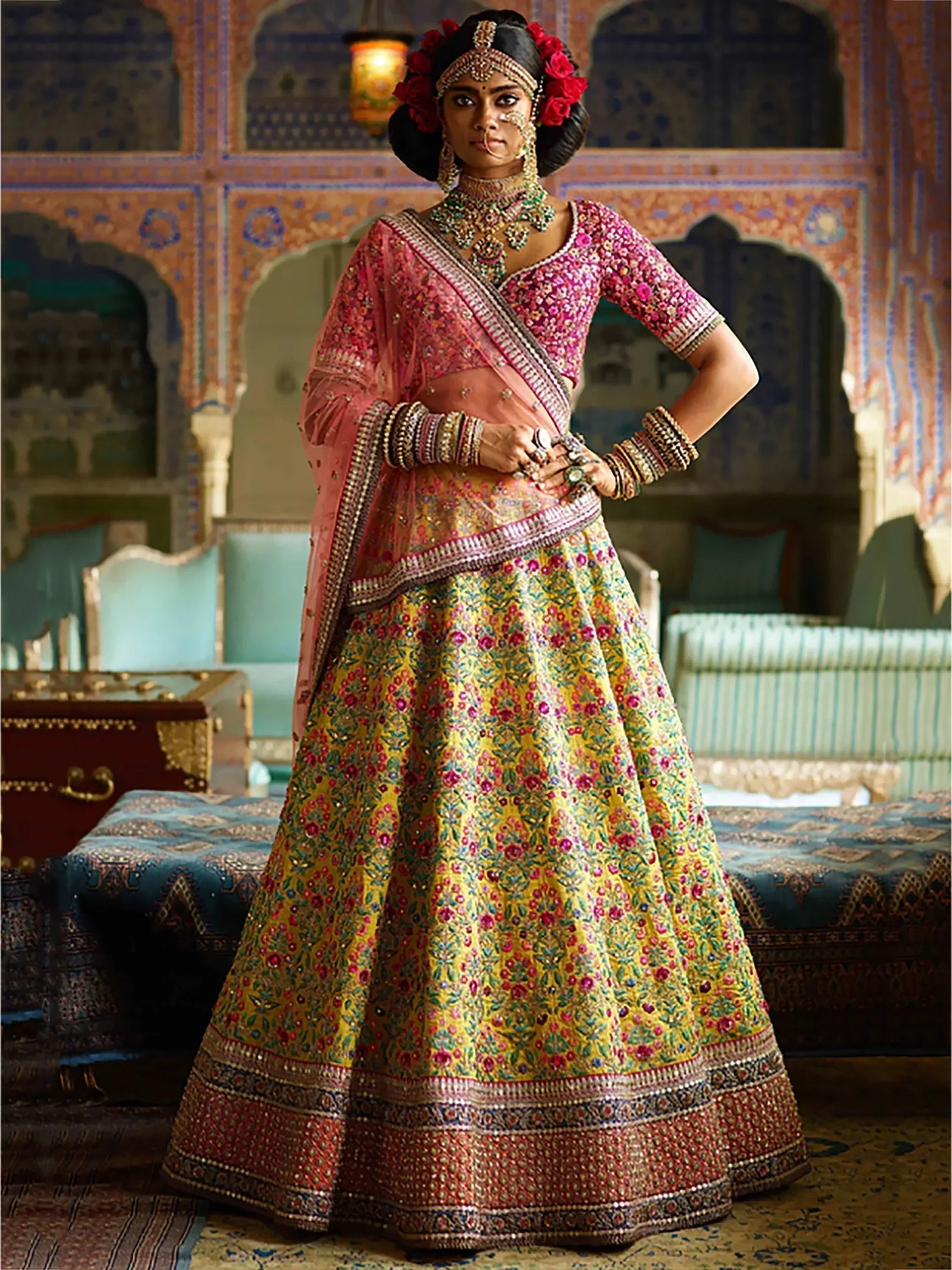 Indian bride in vibrant floral lehenga choli with heavy embroidery, traditional jewelry, and red floral hair accessories